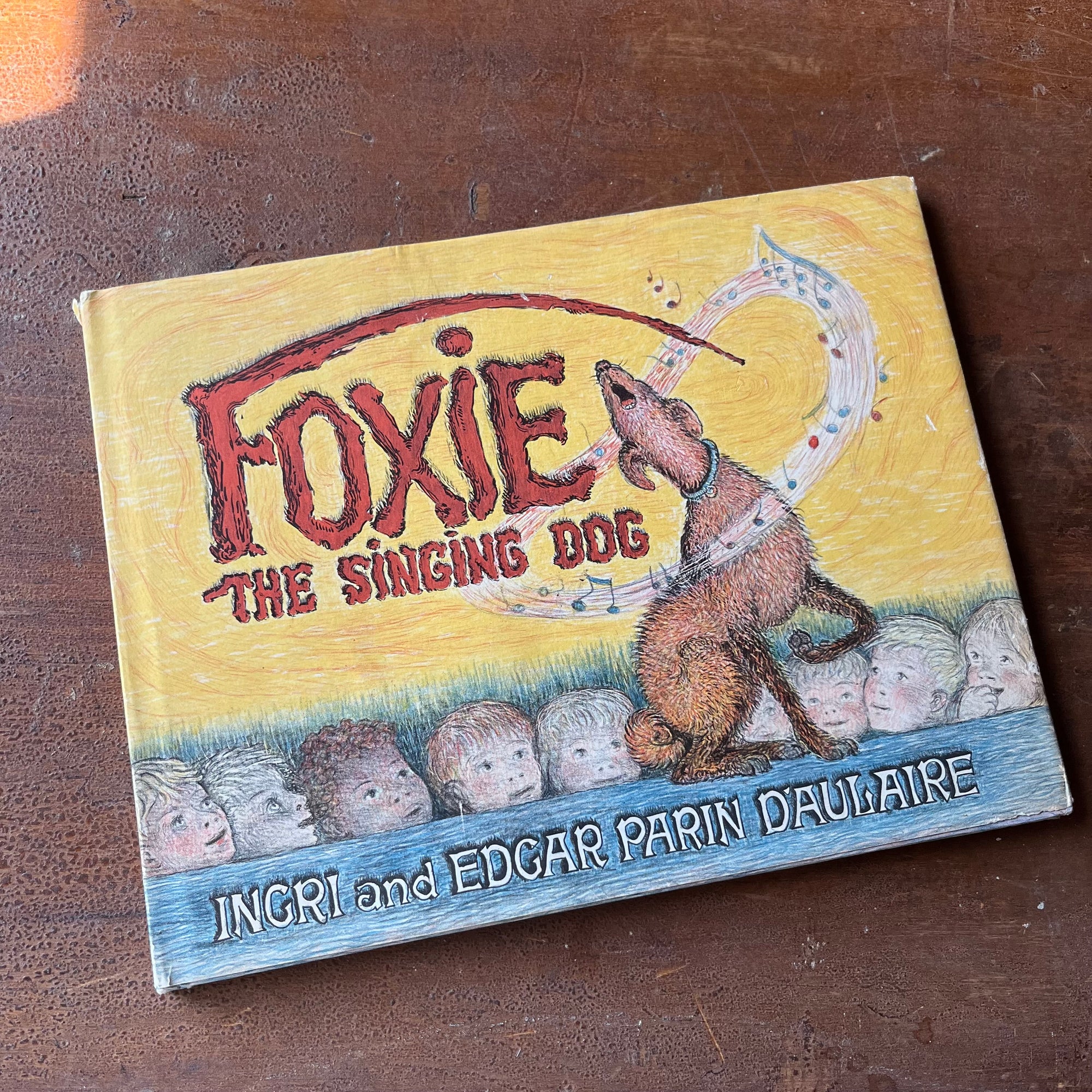 vintage children's picture book - Foxie the Singing Dog written and illustrated by Ingri and Edgar Parin D'Aulaire - a hard cover 1969 edition with dust jacket - view of the dust jacket's front cover with foxie the dog singing with a line of children watching him sing