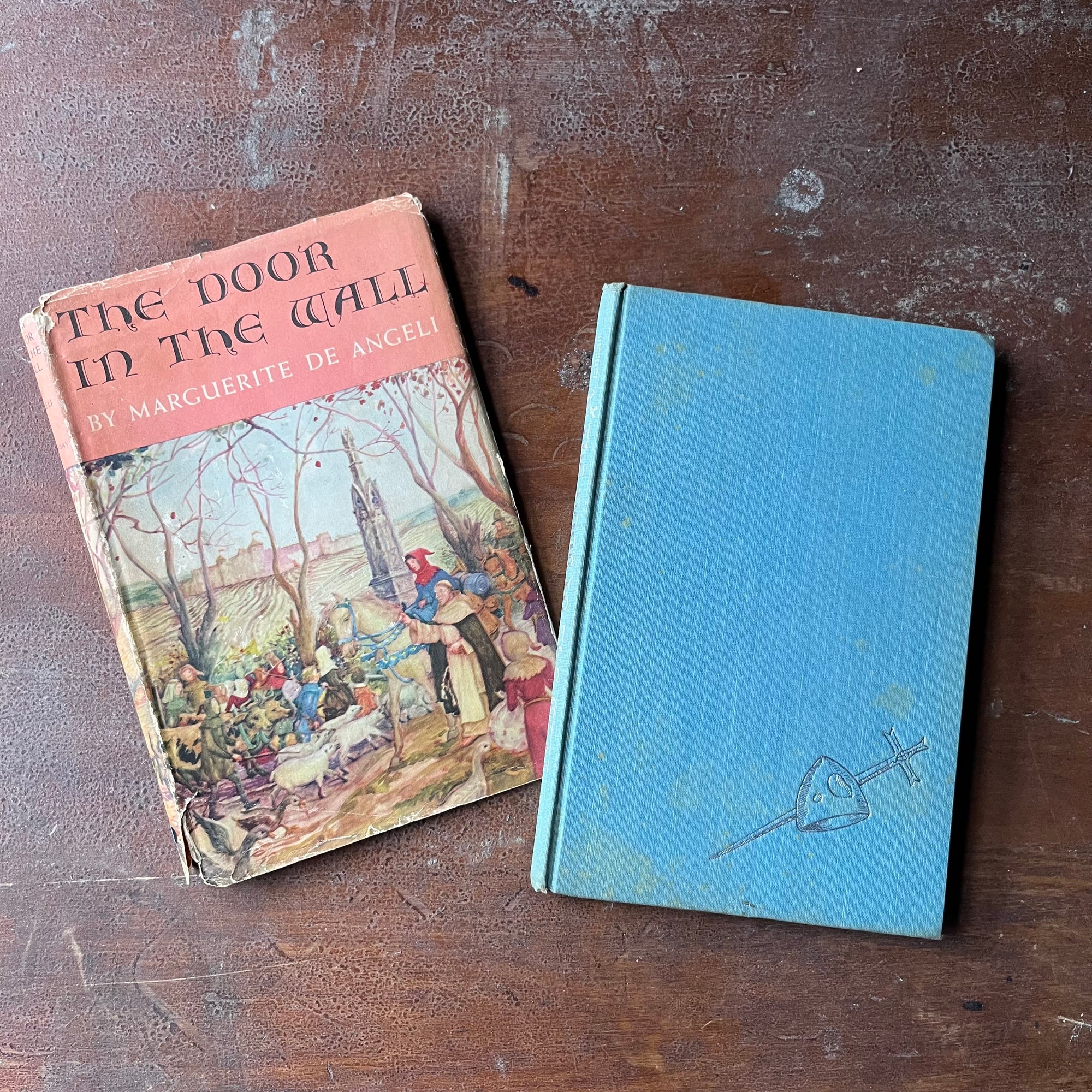 The Door in the Wall by Margeurite de Angeli-1949 Stated First Edition with Dust Jacket-vintage John C. Newbery Medal Winner - view of the dust jacket's front cover along with the book's front cover - the book is blue with an embossed sword stuck through what I think is a helmet? on the bottom right hand corner