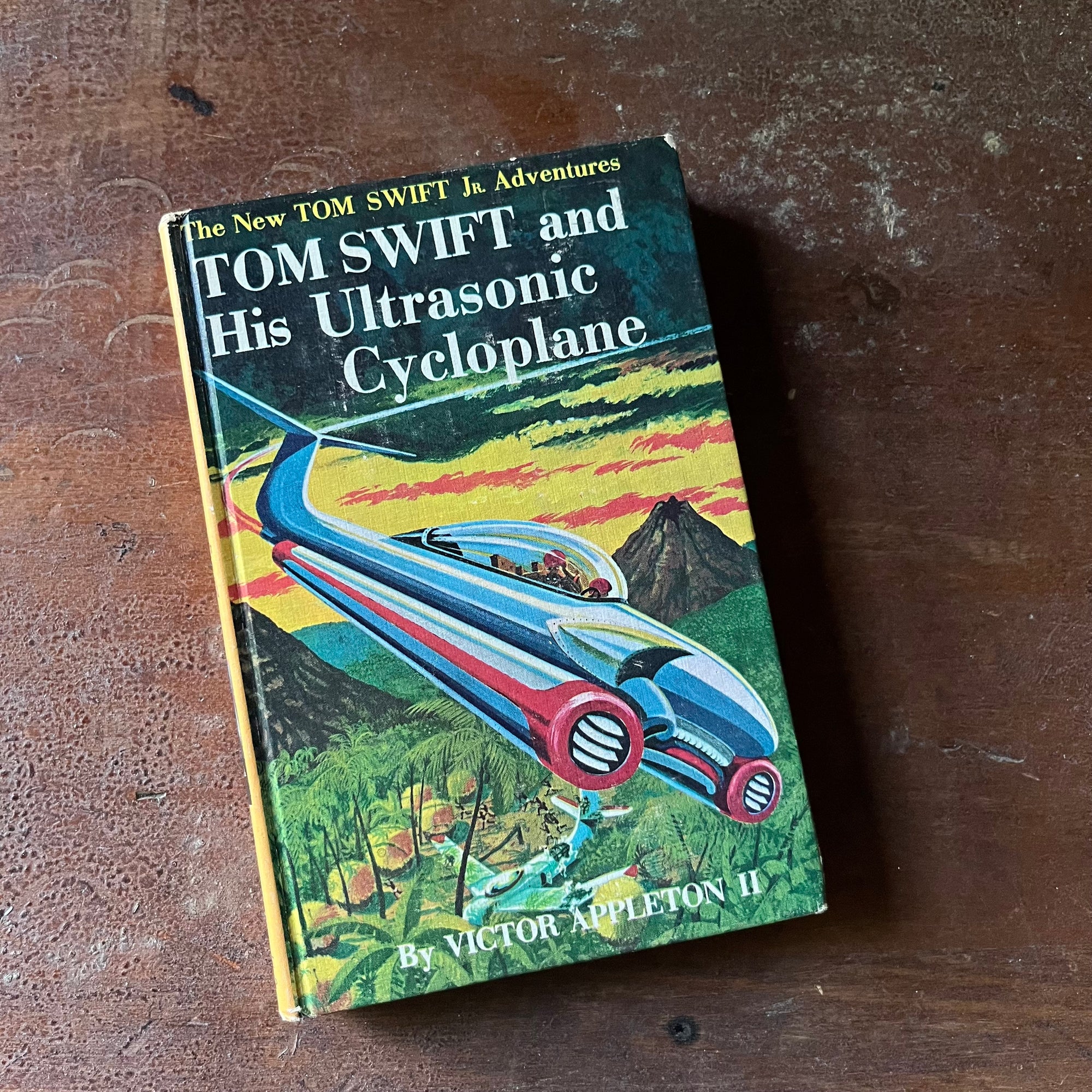 vintage children's chapter book, The New Tom Swift Jr. Adventure Book - Tom Swift and His Ultrasonic Cycloplane written by Victor Appleton II with illustrations by Graham Kaye - view of the front cover in vivid colors showing the cycloplane zooming above a jungle landscape