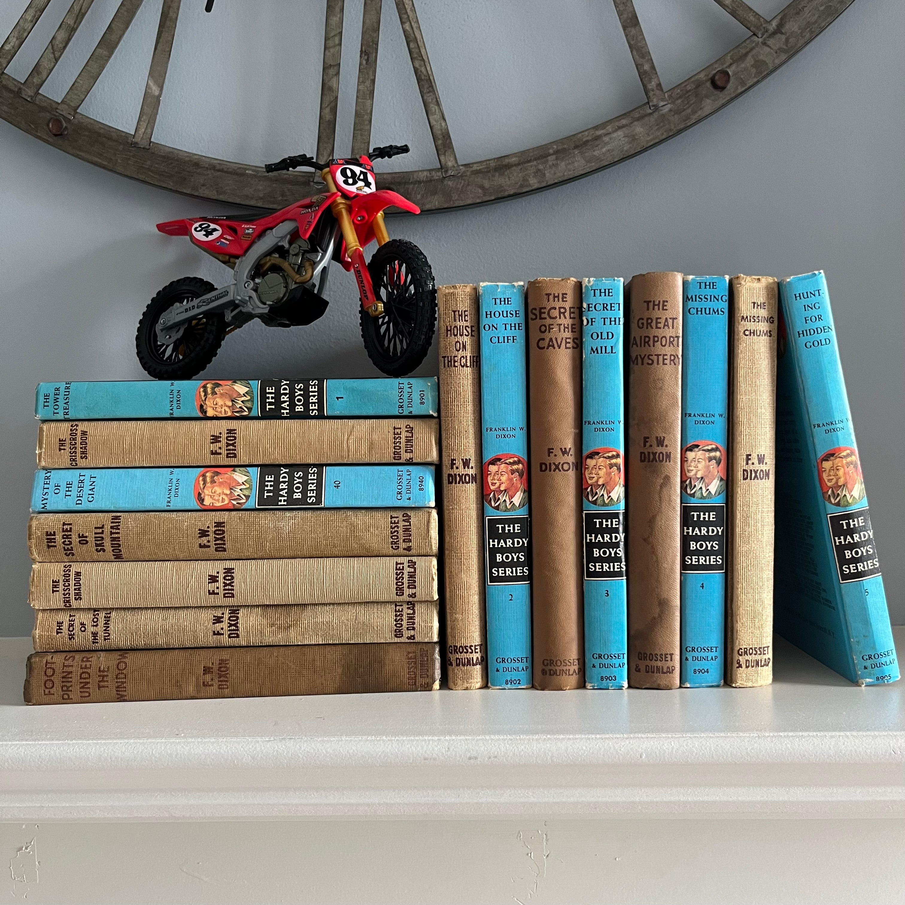 a collection of antique & vintage Hardy Boys books stacked on a mantle with a plastic dirt bike sitting on top of them