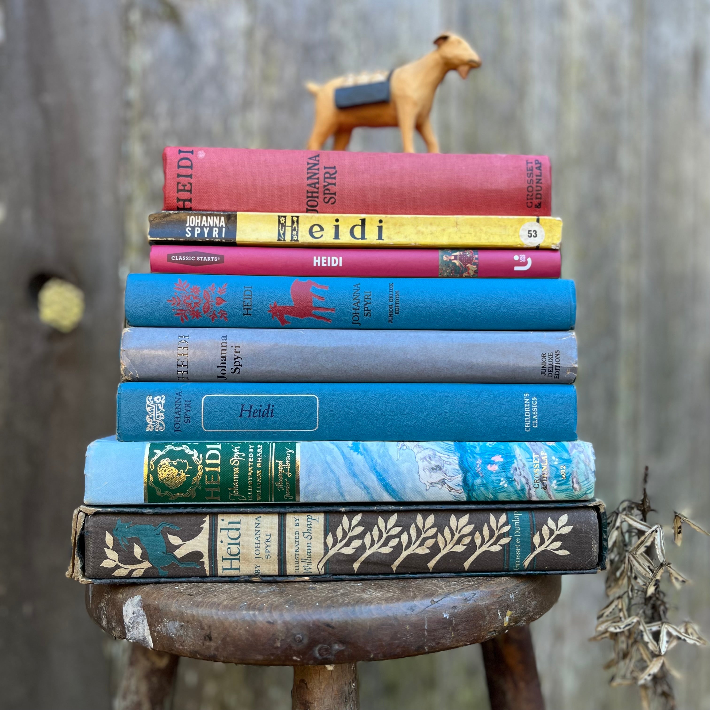 a stack of vintage Heidi books by Johanna Spyri sitting on a stool with a wooden goat perched on top of the book stack