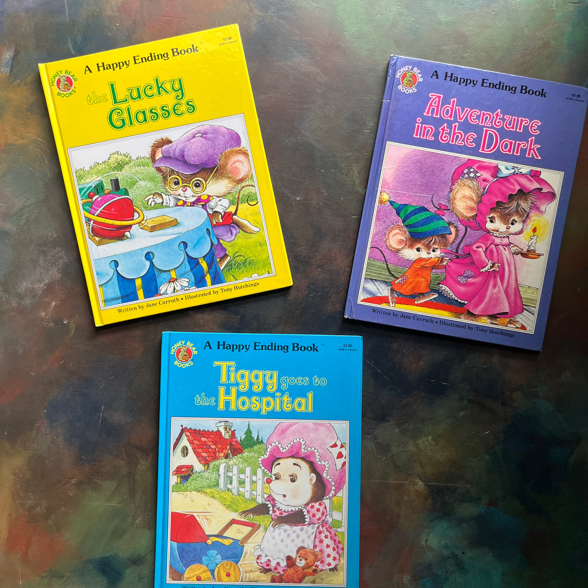 Three vintage children's books from the 'A Happy Ending Book' series written by Jane Carruth with illustrations by Tony Hutchings are arranged on a dark, textured metallic surface. The books feature colorful covers with illustrations of cartoon mice in various adventures: 'The Lucky Glasses' in yellow, 'Adventure in the Dark' in purple, and 'Tiggy Goes to the Hospital' in blue.