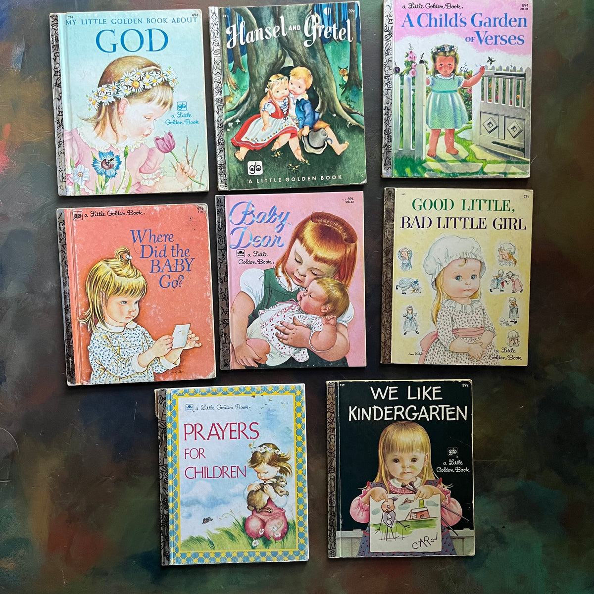 A set of eight vintage Little Golden Books children's picture books arranged in a 3-by-3 grid on a dark, reflective surface. The books feature colorful, classic illustrations and titles including Book about God, Hansel and Gretel, A Child's Garden and Verses, Where Did the Baby Go?, Baby Dear, Good Little, Bad Little Girl, Prayers for Children, and We Like Kindergarten. View of the front covers.