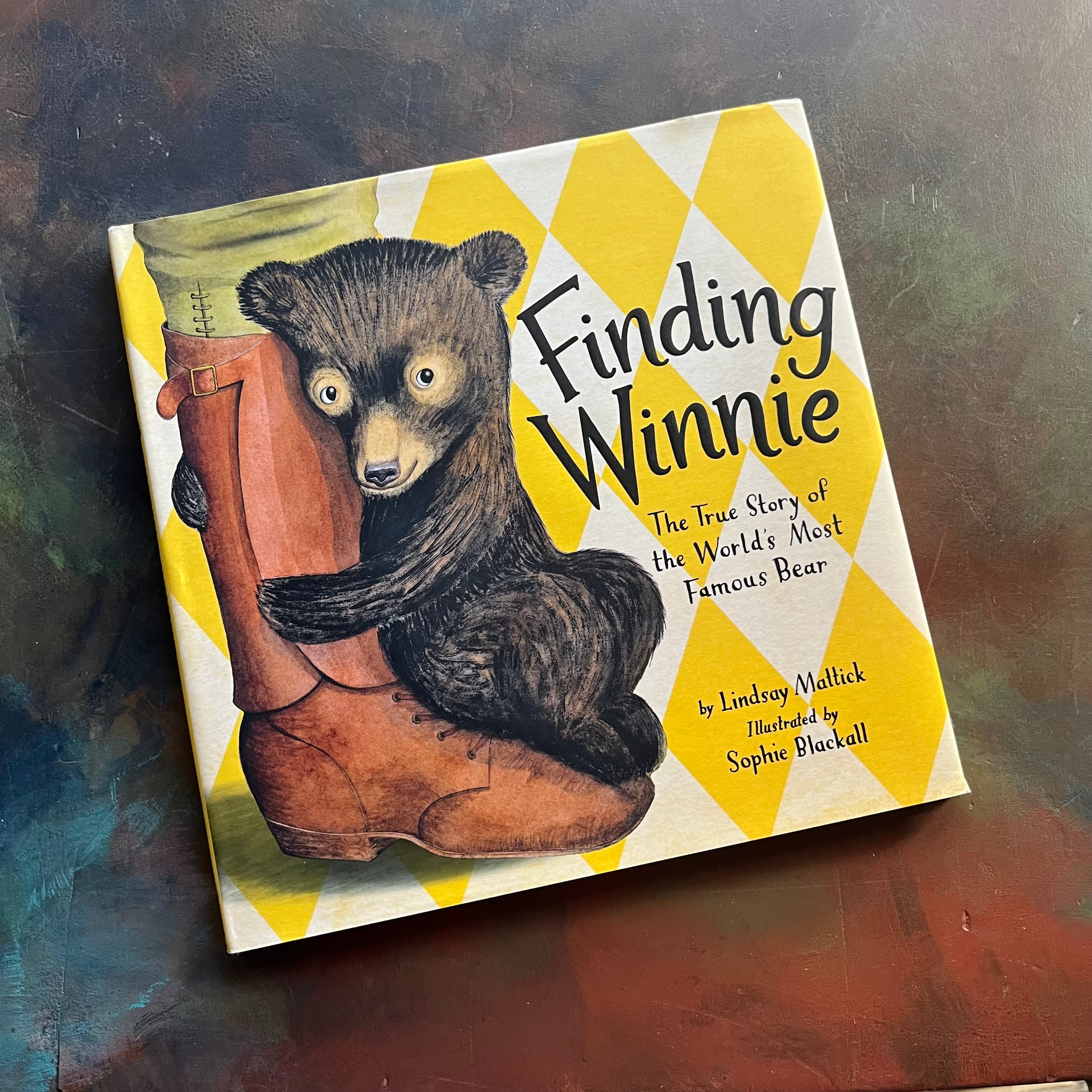 Finding Winnie The True Story of the World's Most Famous Bear written by Lindsay Mattick with illustrations by award winning artist Sophie Blackall-2016 Caldecott Winner-read aloud revival-children's story-Winnie the Pooh-view of the dust jacket's front cover with an image of the bear hugging a soldier's boot