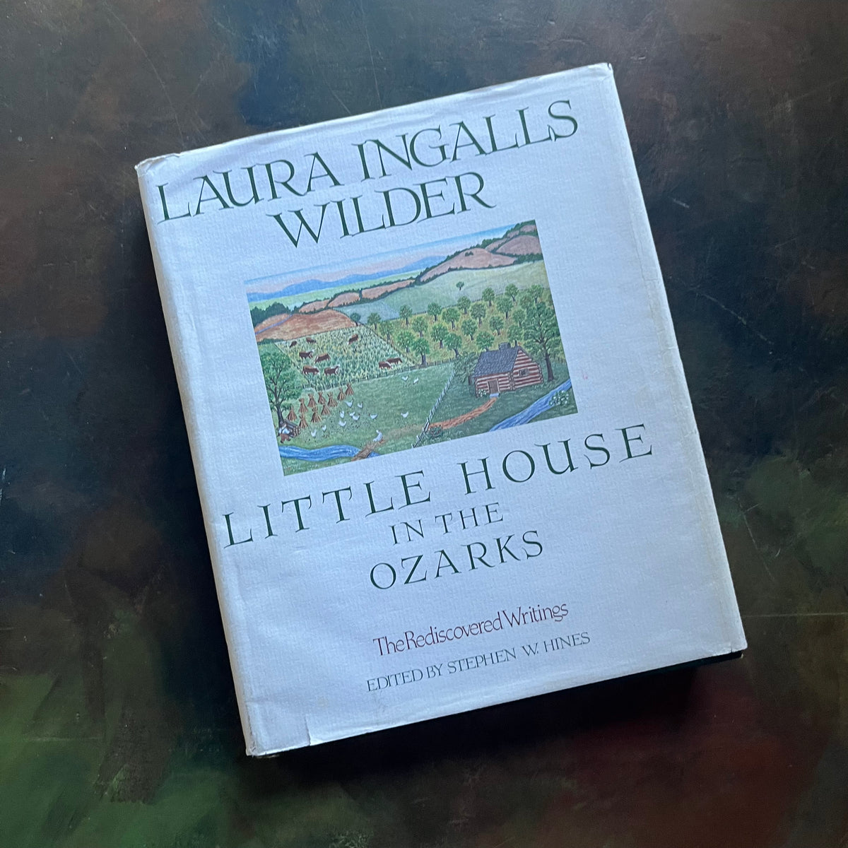 Little House in the Ozarks A Laura Ingalls Wilder Sampler The Rediscovered Writings-Edited by Stephen W. Hines-view of the dust jacket's front cover with a painting depicting a log cabin, a stream, fields, cattle, an orchard and mountains in the background