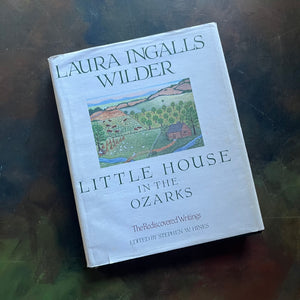 Little House in the Ozarks A Laura Ingalls Wilder Sampler The Rediscovered Writings-Edited by Stephen W. Hines-view of the dust jacket's front cover with a painting depicting a log cabin, a stream, fields, cattle, an orchard and mountains in the background