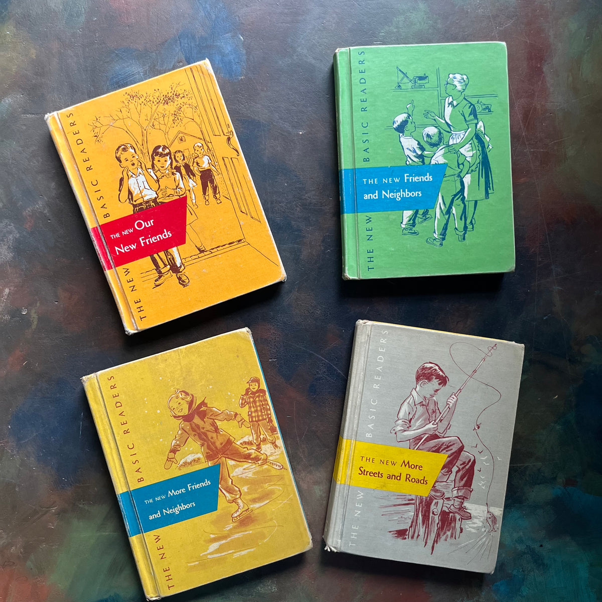 Four vintage hardcover children's books arranged on a dark textured surface. The books include 'The Old Our New Friends' in orange with a red band, 'The New Friends and Neighbors' in green with a blue band, 'The New More Friends and Neighbors' in yellow with a blue band, and 'The New More Streets and Roads' in gray with a yellow band. New Basic Readers published by Scott, Foresman and Company Publishers as part of the Curriculum Foundation Series. 
