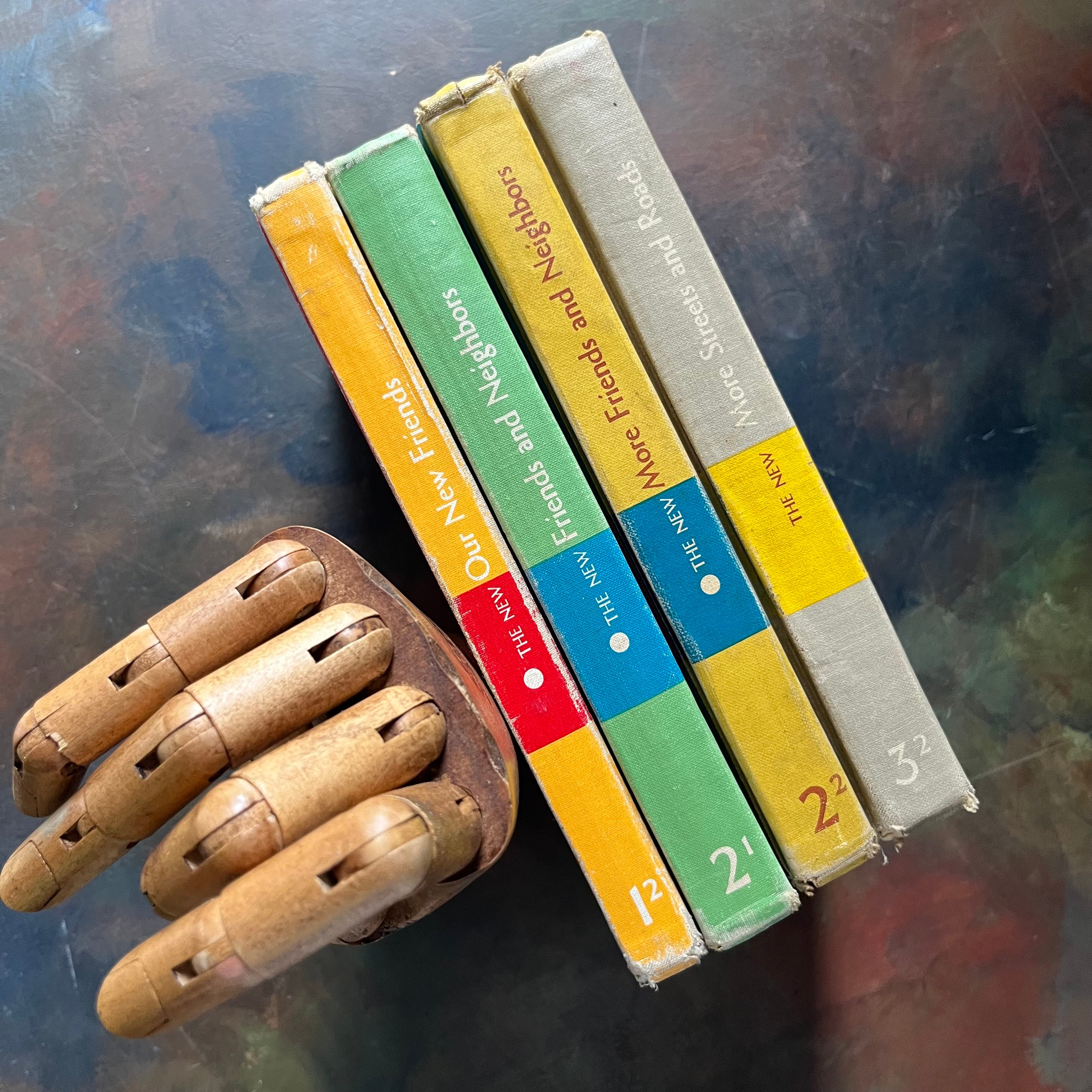 Four vintage children's books from the New Basic Readers published by Scott, Foresman and Company Publishers as part of the Curriculum Foundation Series, displayed vertically (showing their spines) next to a wooden articulated hand on a dark, textured surface.