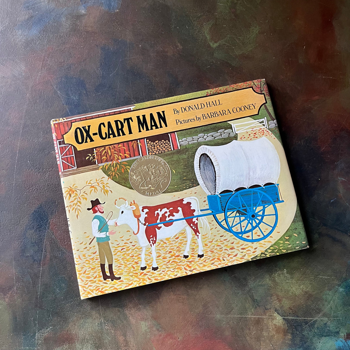 Ox-Cart Man written by Donald Hall with illustrations by Barbara Cooney-vintage children's picture book-1980 Caldecott Medal Winner-view of the book's front cover featuring an illustration of a man with his ox and cart or covered wagon.