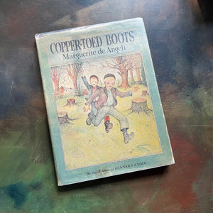 The Copper-Toed Boots written and illustrated by Marguerite de Angeli-first edition-vintage children's biography-chapter book-view of the dust jacket's front cover with an image of two little boys running through a pasture with tree stumps, a fence, a house and barn visible as well as grown trees