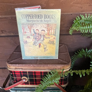 The Copper-Toed Boots written and illustrated by Marguerite de Angeli-first edition-vintage children's biography-chapter book-view of the book sitting on an old plaid picnic basket in front of a log cabin wall with a Boston fern on the right hand of the image