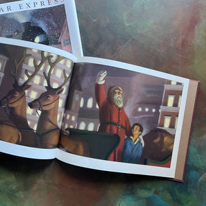 The Polar Express written and illustrated by Chris Van Allsburg-Caldecott award winning picture book-Christmas classic-view of the illustration of Santa gifting the first gift of Christmas, a sleigh bell to the boy with reindeer and the North Pole buildings in the background
