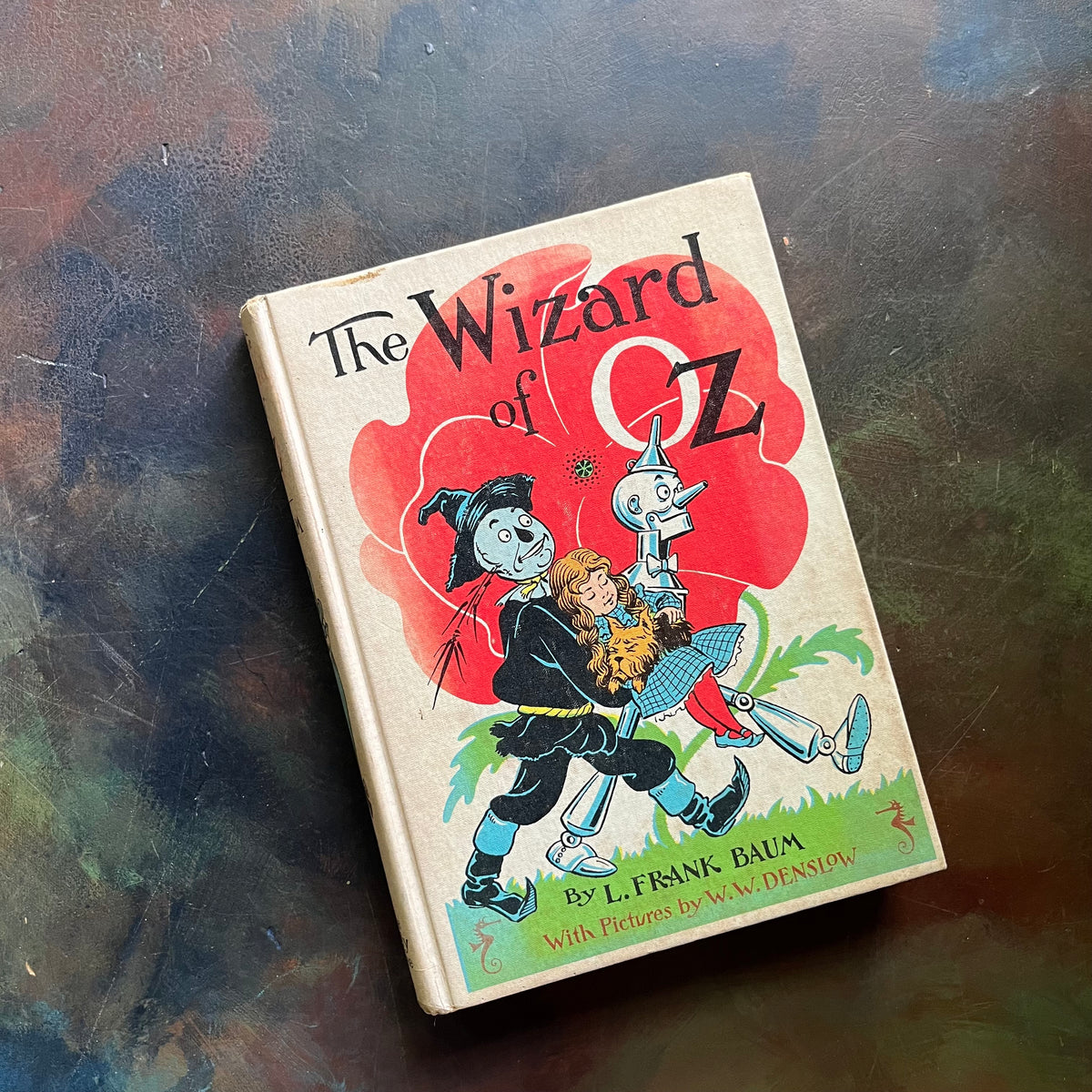 The Wizard of Oz by written by L. Frank Baum with illustrations by W. W. Denslow-1956 Edition-modern day fairy tale-classic children's literature-view of the clothbound front cover with an illustration of the scarecrow and the tinman carrying a sleeping Dorothy while she is holding Toto. 