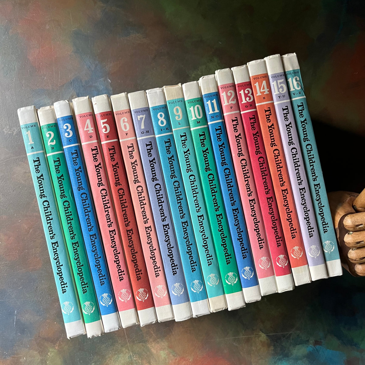 Row of books titled 'The Young Children's Encyclopedia' held by a hand on a textured surface. The Young Children's Encyclopedia Complete 16 Volume Set-Encyclopedia Britannica-1970's living kids books-living books-view of the spines