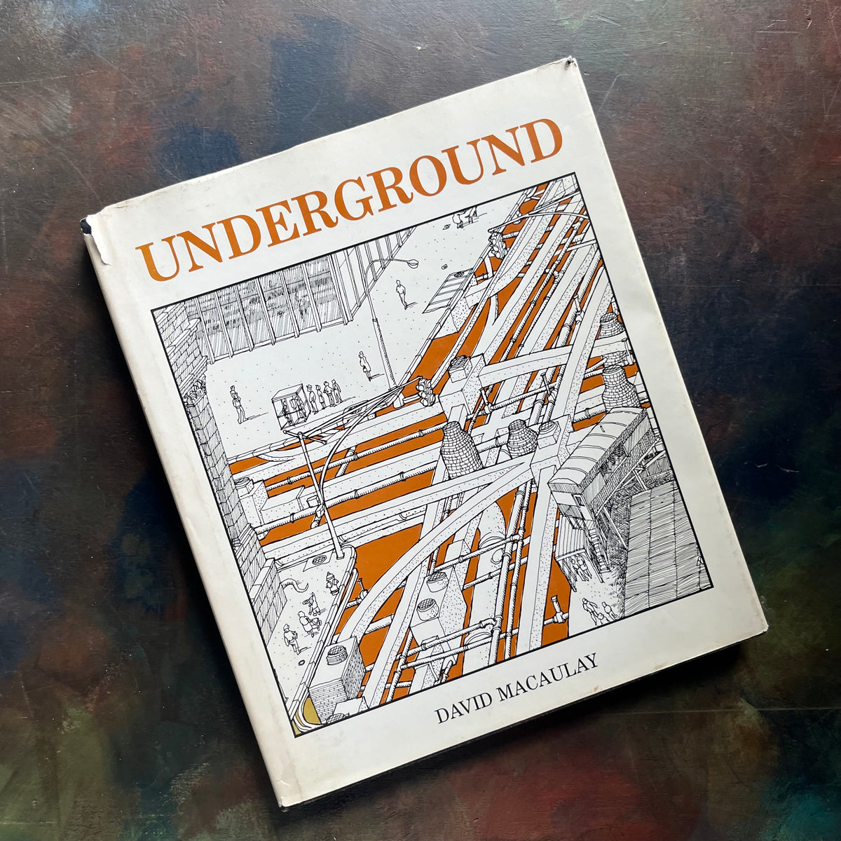 A hardcover art book titled 'UNDERGROUND' by David Macaulay, featuring a black and white architectural illustration with orange accents on the cover, photographed on a dark textured surface.