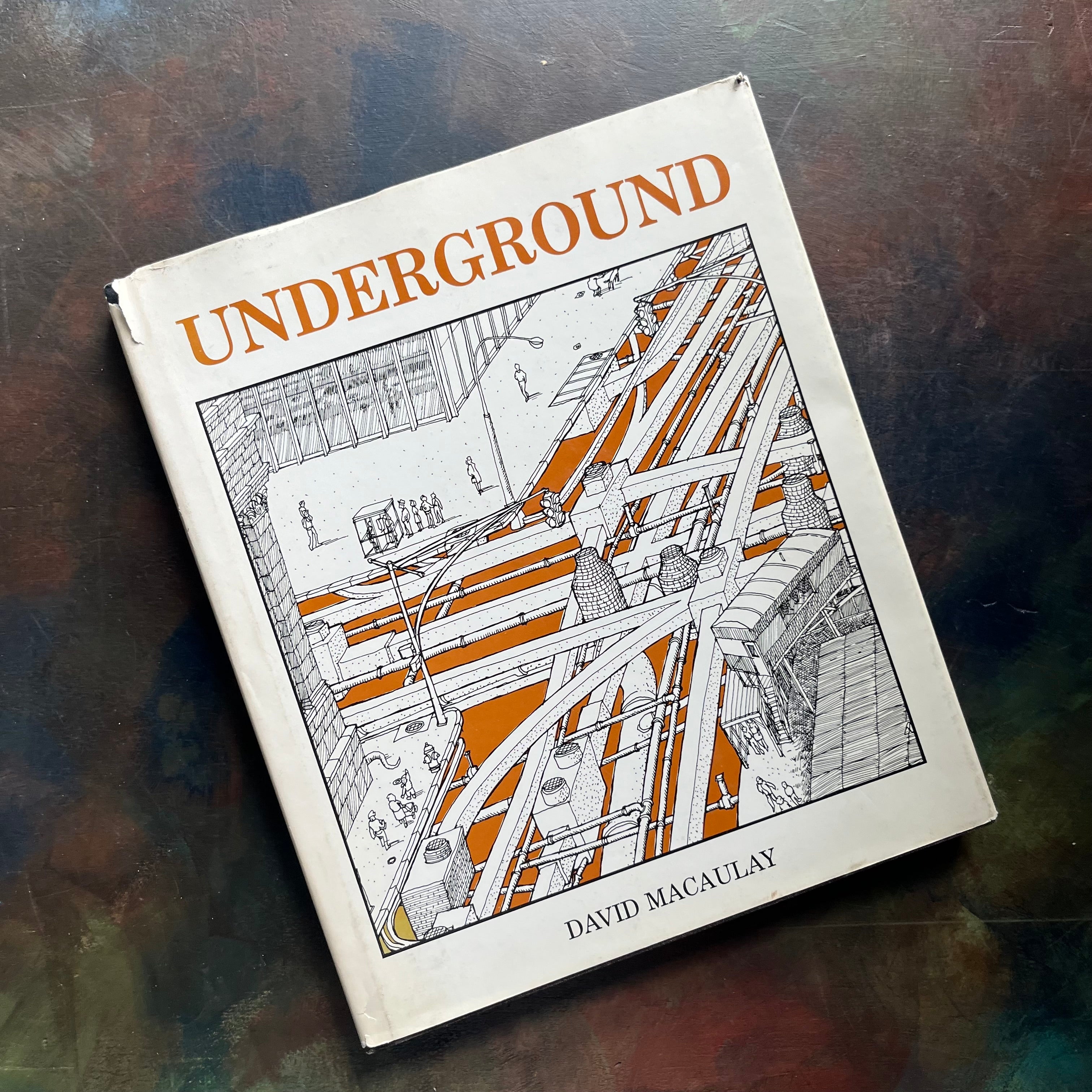 A hardcover art book titled 'UNDERGROUND' by David Macaulay, featuring a black and white architectural illustration with orange accents on the cover, photographed on a dark textured surface.