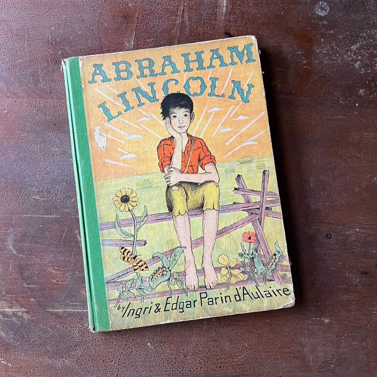 Log Cabin Vintage – vintage children’s book, children’s book, chapter book, living history book, history book, non-fiction, picture book - Abraham Lincoln by Ingri & Edgar Parin d'Aulaire - view of the front cover with an illustration of Lincoln sitting on a split rail fence as a child